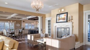 A spacious living room with a fireplace, a dining area, and a kitchen in the background. - Luxurious 6 bedroom estate with lake view & beach (Fennville)