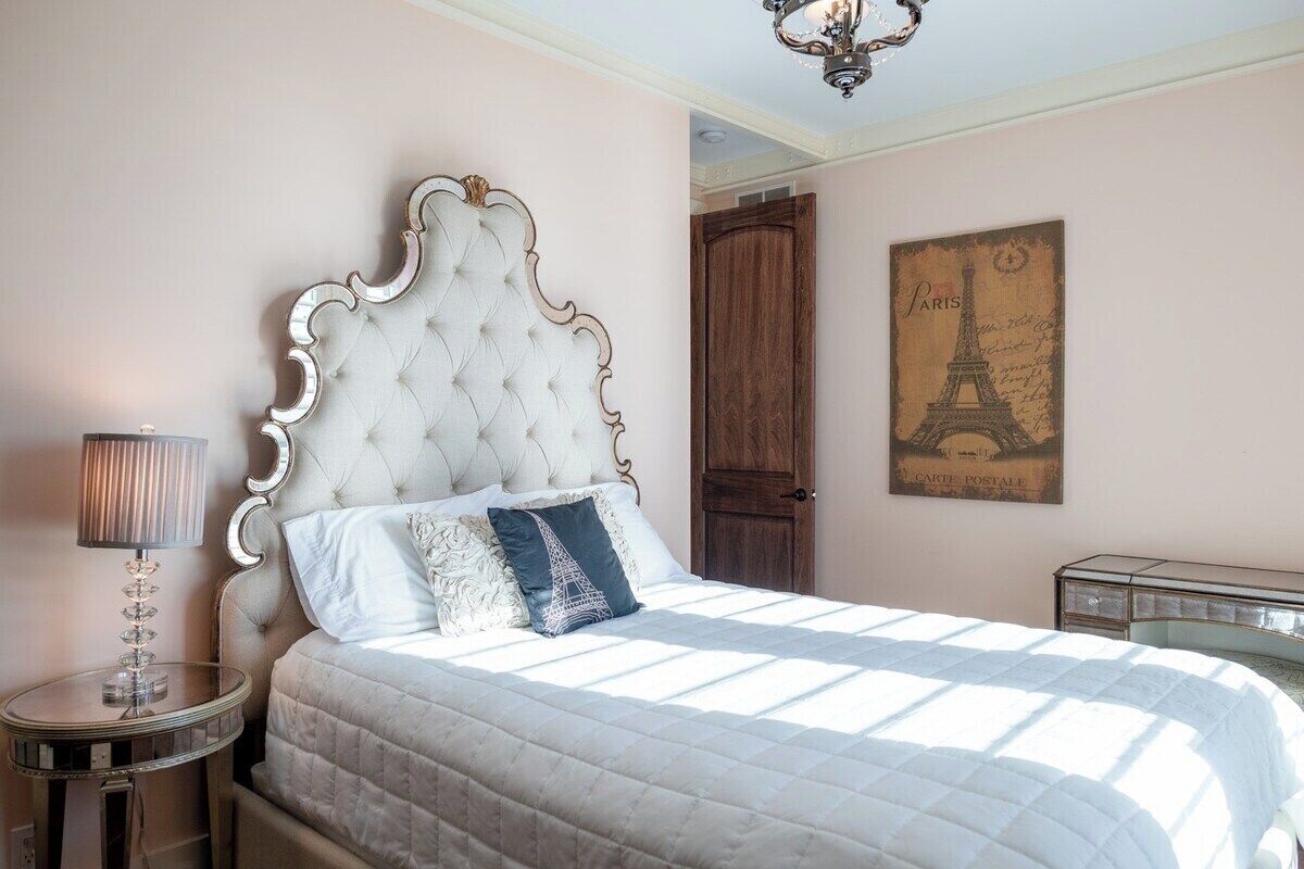 Un dormitorio con cabecera capitonada, cama con cubrecama acolchado, lámpara de noche, puerta de madera y una pared decorada con un cartel con temática de París.