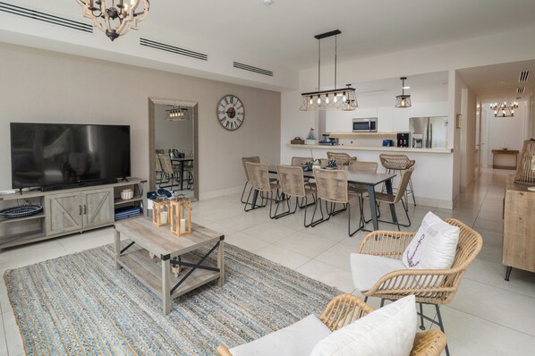 A modern living room with a flat-screen TV, wicker furniture, and a dining area with a kitchen in the background.