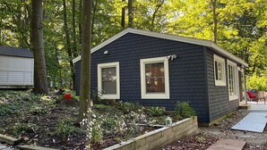 A small, dark-colored house with two windows, surrounded by a garden with various plants and a wooden deck. - Updated Cozy Cabin near Lake Michigan (Holland)