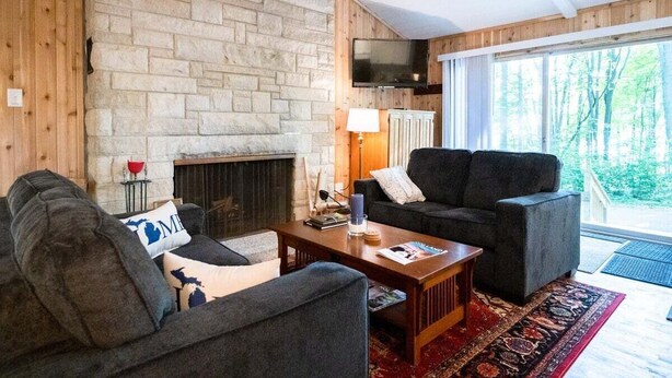 A cozy living room with a stone fireplace, two grey sofas, a wooden coffee table, and a large window with a view of trees. - Updated Cozy Cabin near Lake Michigan (Holland)