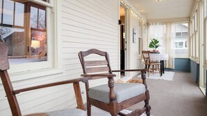 A porch with wooden chairs and a table, white siding walls, and a view of a house through the windows.