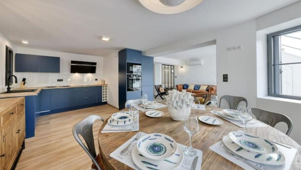 Una cocina moderna con armarios azules, un comedor con mesa de madera dispuesta para seis personas y una zona de estar acogedora con sofá y sillones.