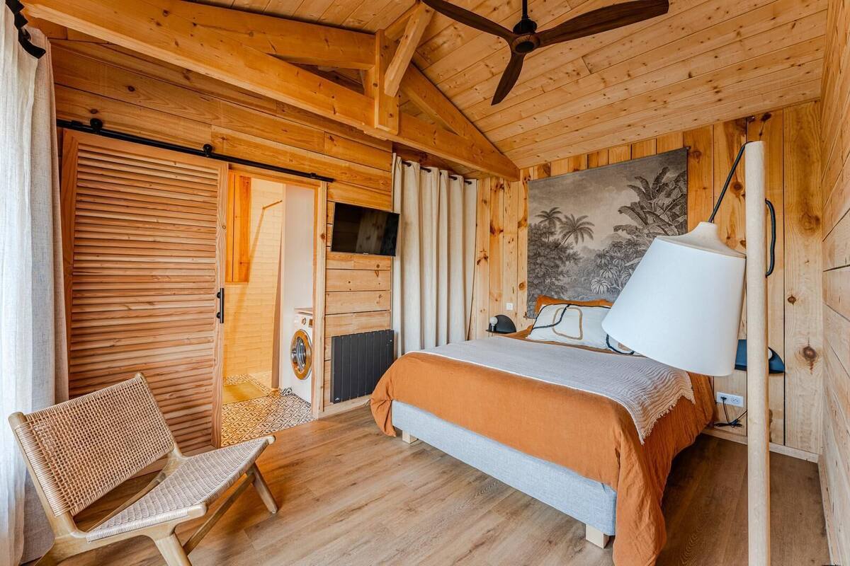 A wooden room with a bed, a chair, a ceiling fan, and a wall with a tropical mural.