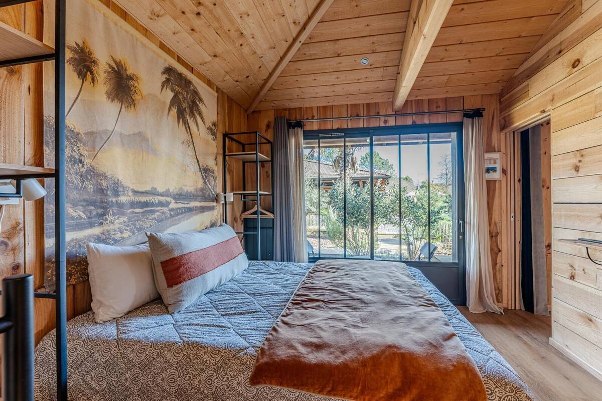 A wooden cabin room with a bed, a large window, and a mural of palm trees.