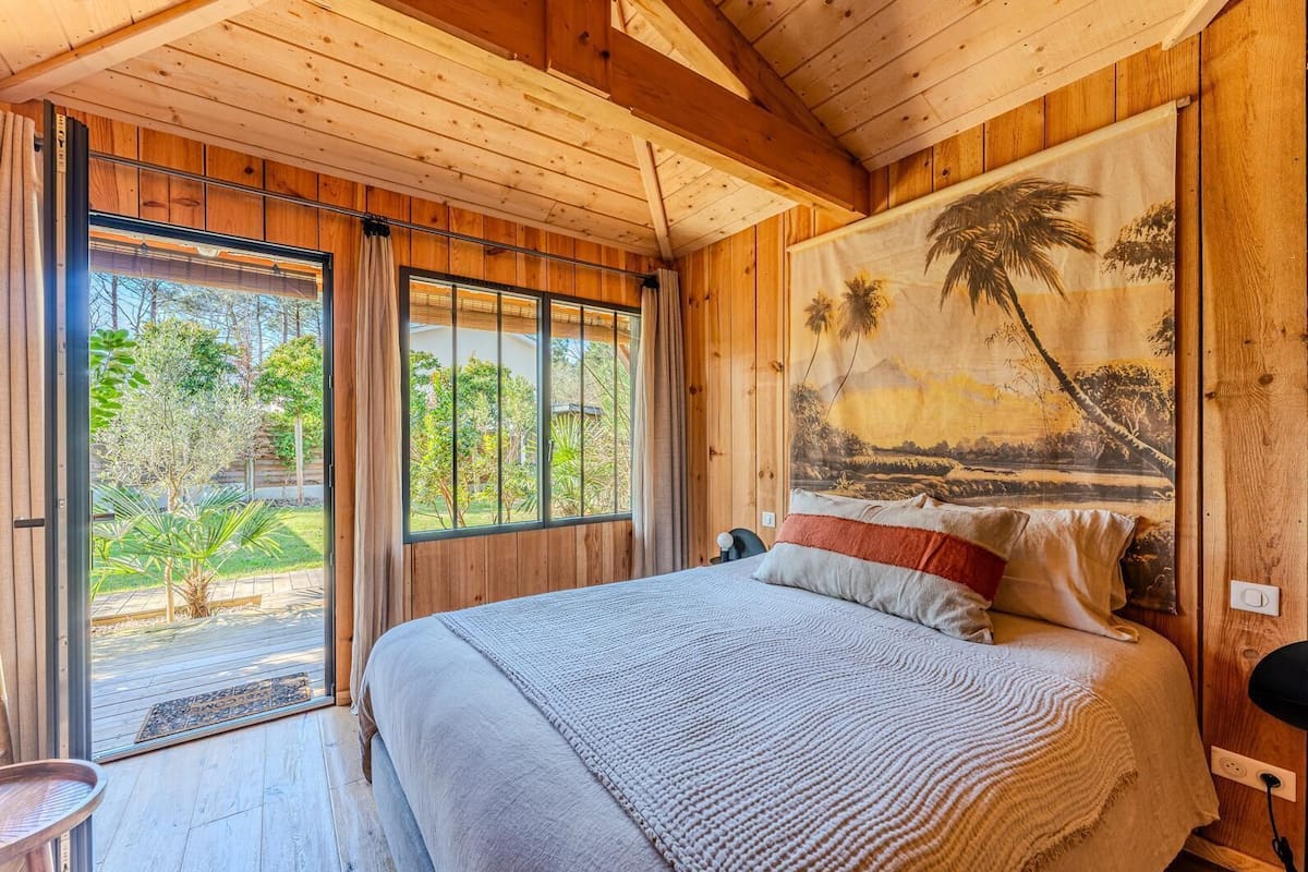 A wooden room with a bed, a large window, and a tropical mural.