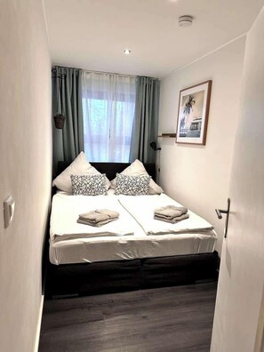 A double bed with patterned pillows, a small bedside table, a framed picture on the wall, and a window with curtains.