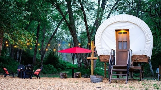 A rustic cabin with a wooden door and a ladder leading up to a platform. There is a red umbrella and a bench with cushions nearby.