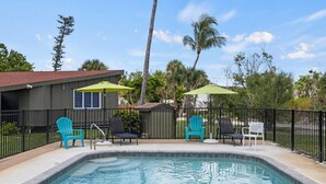 A swimming pool with lounge chairs and umbrellas, surrounded by a fence and palm trees.