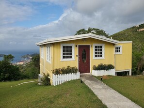 A small yellow building with a red door, white picket fence, and a concrete path leading to the entrance.
