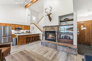 A modern kitchen with a stone fireplace, wooden cabinets, a stainless steel refrigerator, and a bar area.