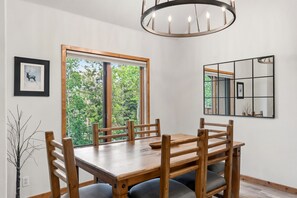 Un'area pranzo con un tavolo e sedie in legno, un'ampia finestra vista alberi, un lampadario e uno specchio sulla parete.