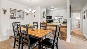 A dining area with a wooden table and chairs, a chandelier, and a kitchen in the background.