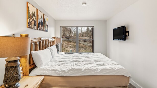 A bedroom with a large bed, a TV mounted on the wall, and a window with a view of trees and hills.