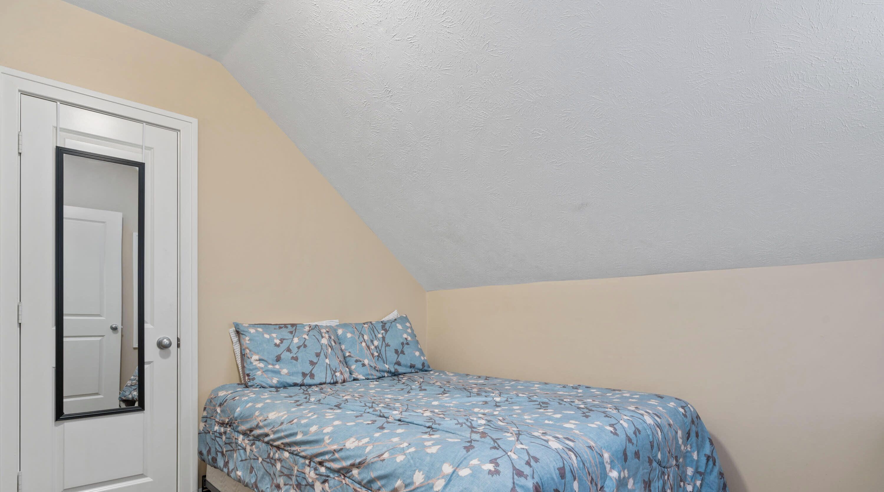 A bedroom with a sloped ceiling, a bed with floral bedding, and a door with a mirror.