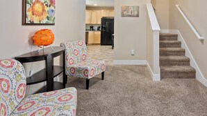 Ein Wohnzimmer mit Sofa, Sessel und Hocker, einem Esstisch, einer Küche im Hintergrund und einer Treppe.
