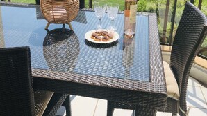 Een tafel met glazen blad, rieten stoelen, een geweven mand, een fles en een bord eten op een balkon met uitzicht op een groen landschap.