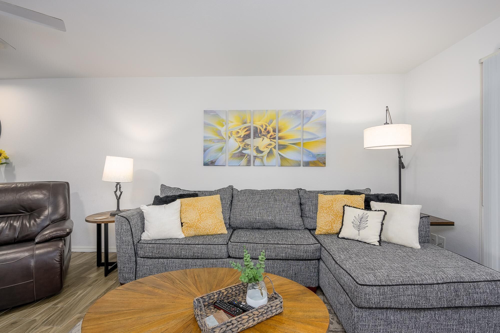 A living room with a grey sectional sofa, a brown leather armchair, a round wooden coffee table, and a floor lamp.