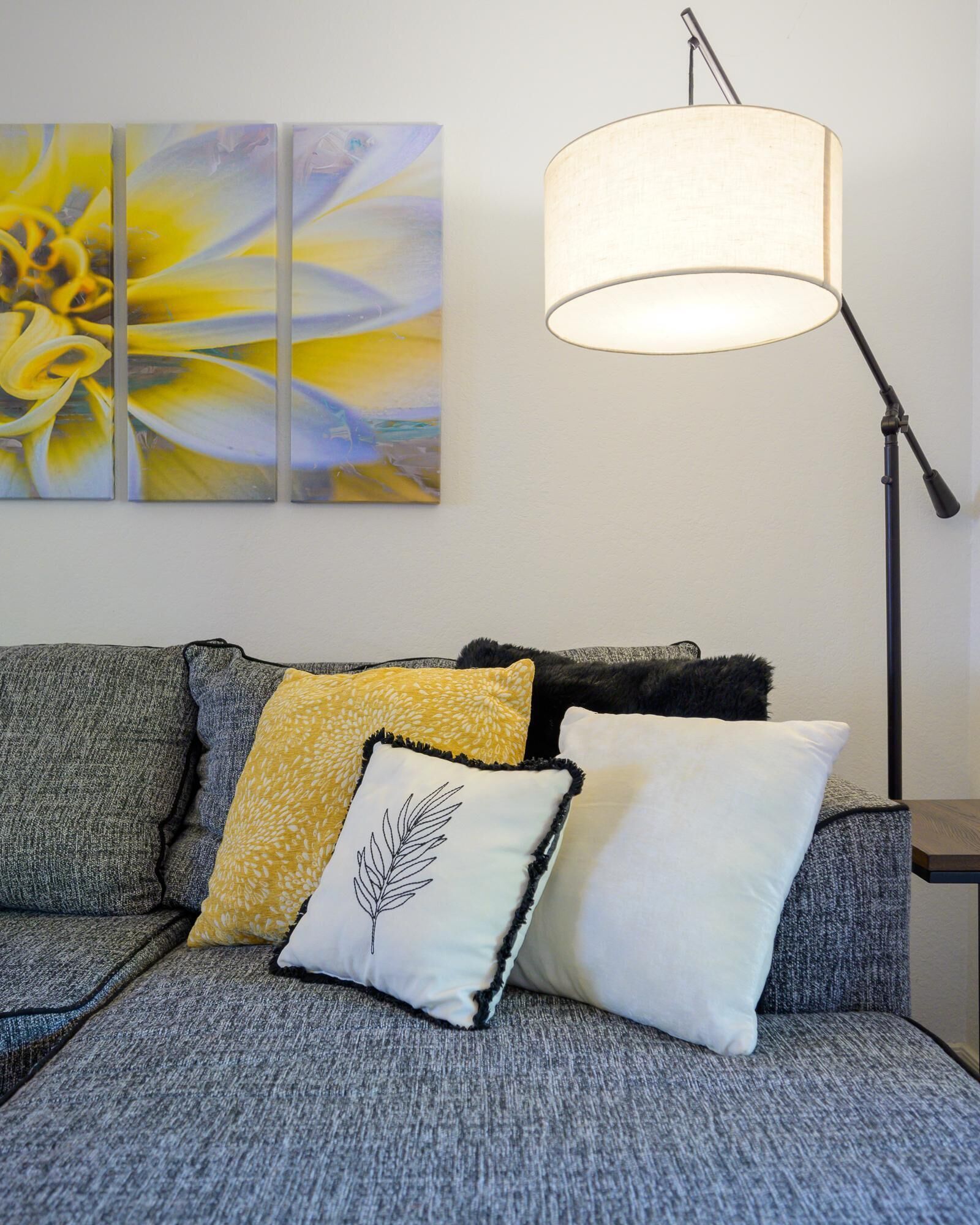 A grey sofa with three pillows, a floor lamp, and a set of three abstract paintings on the wall.