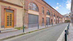 A street with a row of buildings featuring brick facades, a yellow door, and a closed shutter. - LA PARENTHÈSE - Charmant T2 entièrement rénové (Toulouse)