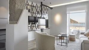 A modern living room with a stone accent wall, a flat-screen TV, a white sofa, a small table, and a view of the beach. - Always Easy I Sagües (San Sebastián/Donostia)