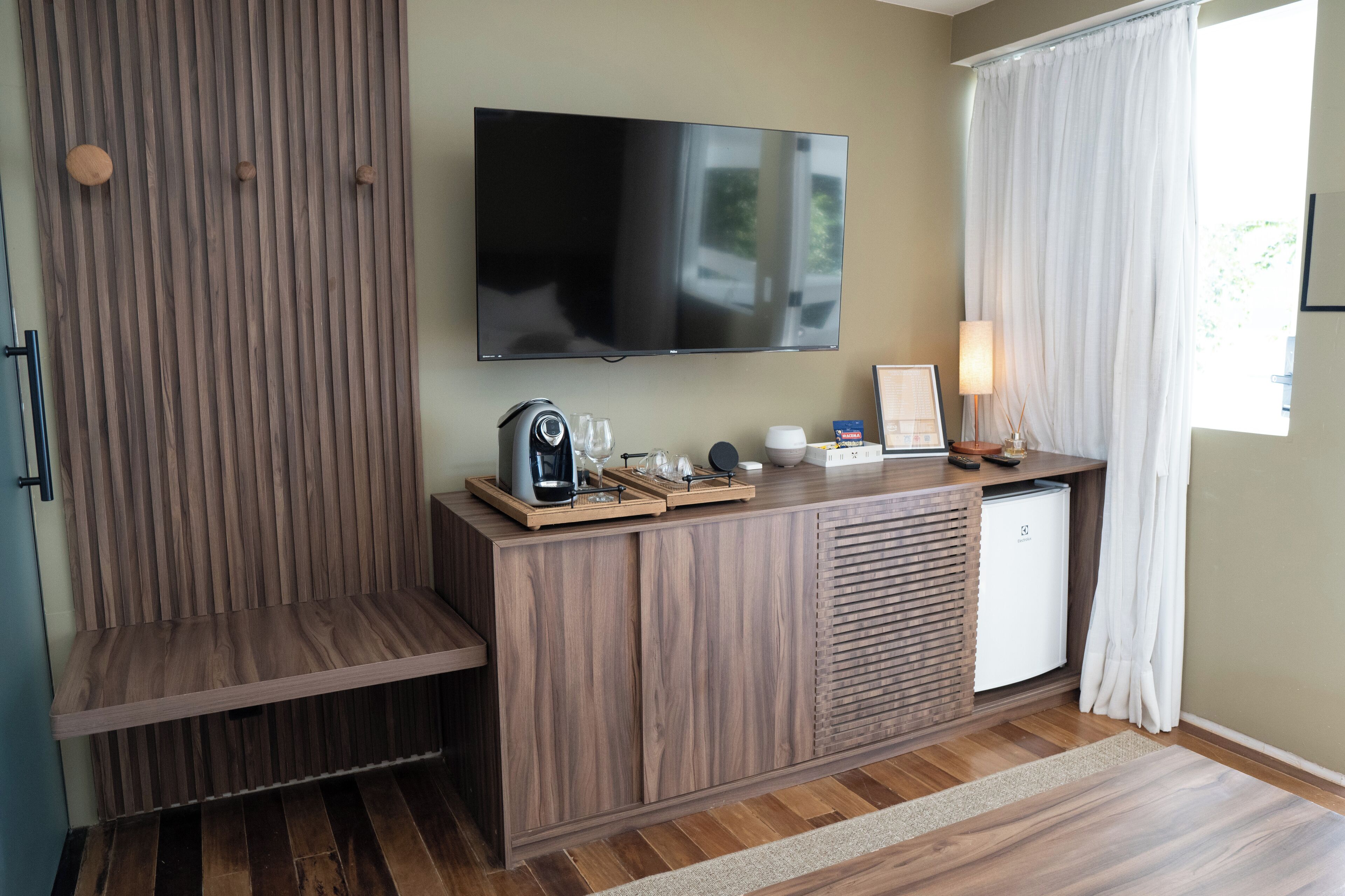Quarto moderno com mesa de console de madeira, TV de tela plana, cafeteira e mini-geladeira.