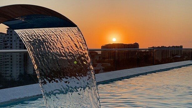 A pool with a waterfall feature, a sunset in the background, and a cityscape. - Entire 2 Bedroom 2 Bath Marina Vallarta condo with insane view  (Puerto Vallarta)