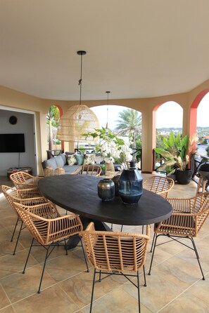 An outdoor dining area with wicker chairs and a round table, surrounded by potted plants and a large woven light fixture.