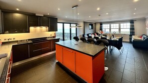 Eine moderne Küche mit dunklen Schränken, einer orangefarbenen Kochinsel und einem Essbereich mit einer schwarzen Ledersofa und einem großen Fenster mit Blick auf die Stadt.