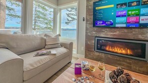 Ein Wohnzimmer mit großem Flachbildfernseher, der Streamingdienste zeigt, einem Kamin, einer Couch und einem Couchtisch mit einem Puzzle und einem Glas Wein.