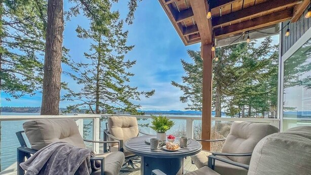 Eine Terrasse mit Außenbestuhlung, einem Tisch mit Pflanze und Getränk sowie Blick auf Wasser und Bäume.