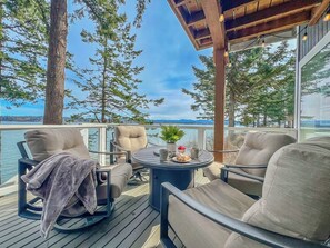 A deck with outdoor seating, a table with a plant and a drink, and a view of the water and trees.