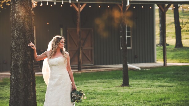 Escena de boda con una novia en vestido blanco sosteniendo un ramo, de pie junto a un árbol con un granero y luces colgantes al fondo.
