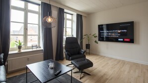Una sala de estar moderna con un ventanal grande, un sillón de cuero negro, una mesita pequeña y un televisor de pantalla plana montado en la pared.