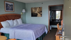 A hotel room with a large bed, a desk, a chair, a lamp, and two paintings on the walls. - Downtown Inn #3- King Room (Asheville)