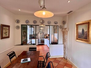 A dining area with a wooden table, chairs, and a book. There is a kitchen visible through a glass door, and a painting on the wall. - La Dommerie - Charming cottage (Domme)