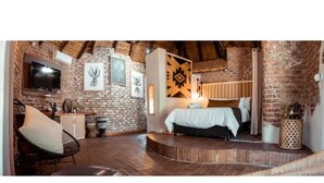 A rustic room with a brick wall, a bed with a white bedspread, a television mounted on the wall, and a wooden floor.