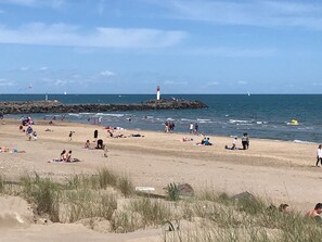 Une plage où des gens se prélassent au soleil et se baignent, un phare en arrière-plan et une digue rocheuse.