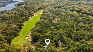 Une vue aérienne d’un terrain de golf entouré d’une forêt dense, avec une étendue d’eau et un ciel dégagé en arrière-plan.