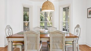Une salle à manger avec une table en bois, des chaises blanches dotées de coussins à rayures, un luminaire suspendu en fibres tressées et de grandes fenêtres donnant sur un paysage verdoyant.