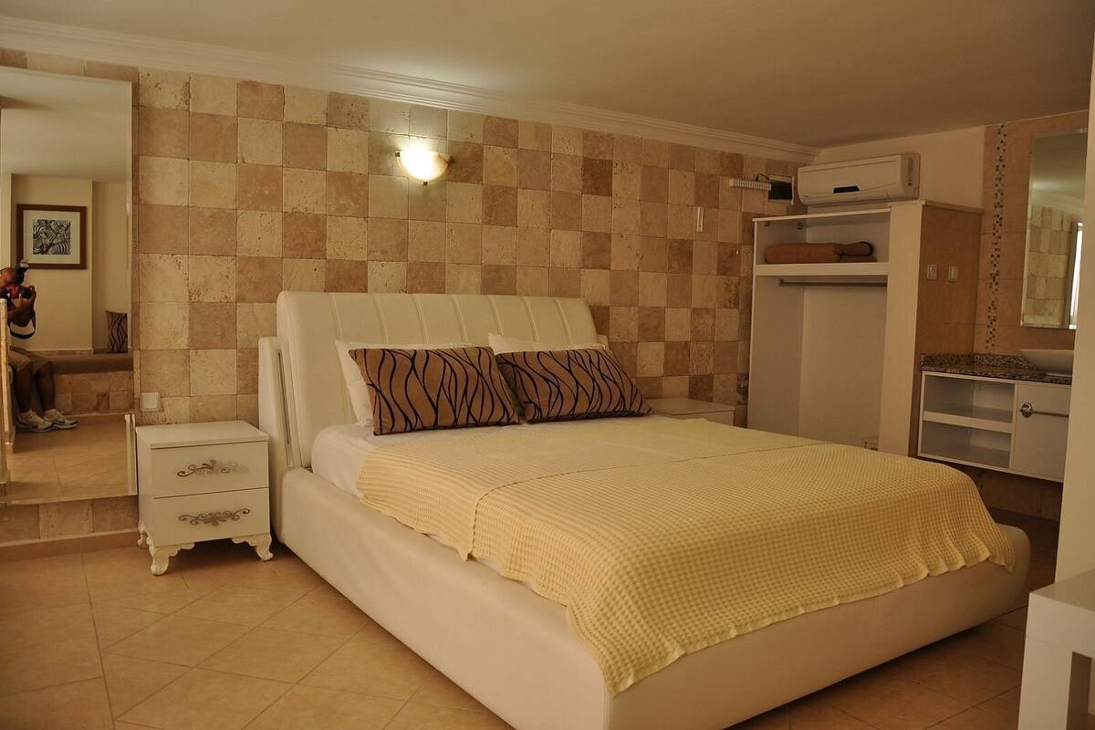 A bedroom with a large bed, a nightstand, a wardrobe, and a bathroom with a sink and mirror.