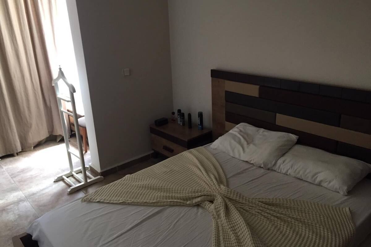 A neatly made bed with a white comforter and pillows, a wooden nightstand with a lamp and a bottle, and a window with curtains.