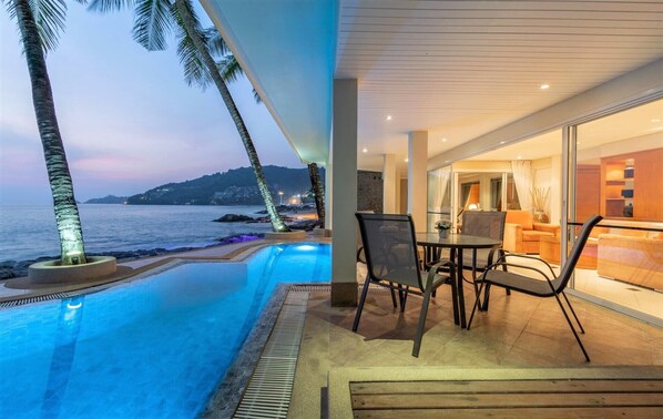 Et moderne hotelværelse med en pool med udsigt over havet, et spiseområde med bord og stole samt udsigt til ocean og bakker.