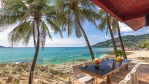 A beachside patio with a table and chairs, overlooking a rocky shoreline and turquoise sea. - Your exclusive villa on the sand, Private Pool, Stunning Ocean Views (Tambon Patong, Amphoe Kathu)