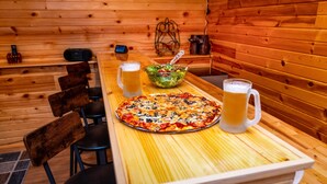 A wooden table with a pizza, a bowl of salad, and two beer mugs in a cabin-style room with wooden walls and chairs. - Covered Hot Tub, Great Games, Near Thunder Bay Falls / With Owners' Club Access (Galena)