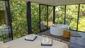 A modern bedroom with a large bed, a bathtub, and a view of lush greenery through large windows. - Peso Village (Amarante)