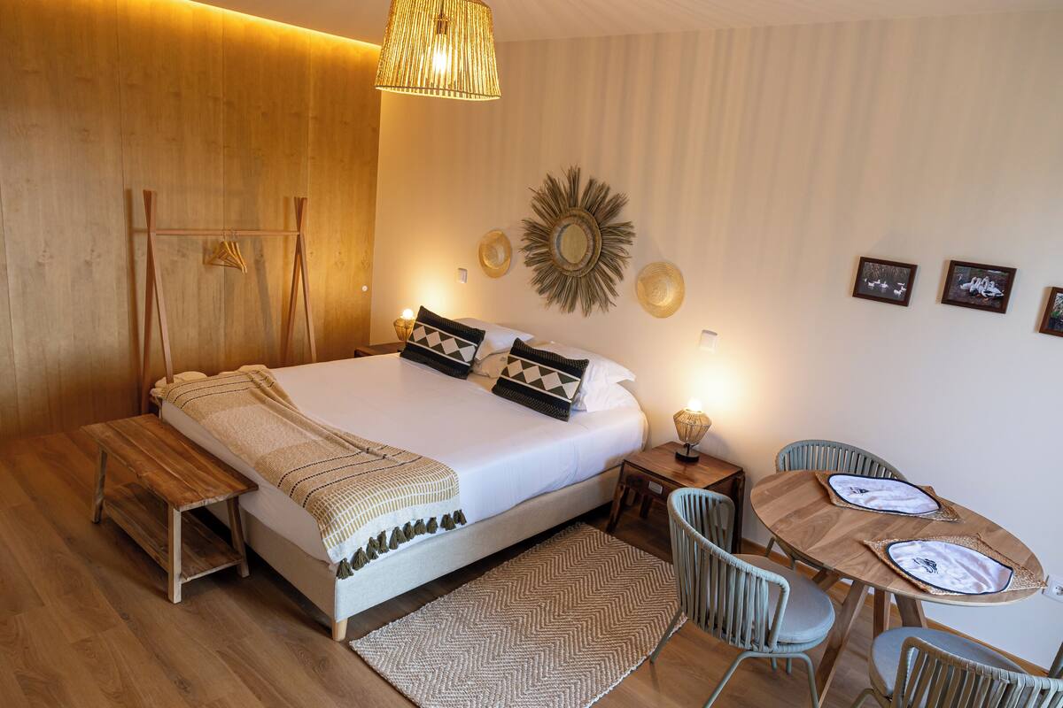 A bedroom with a large bed, bedside tables, a wooden bench, a dining table with chairs, and wall decor.