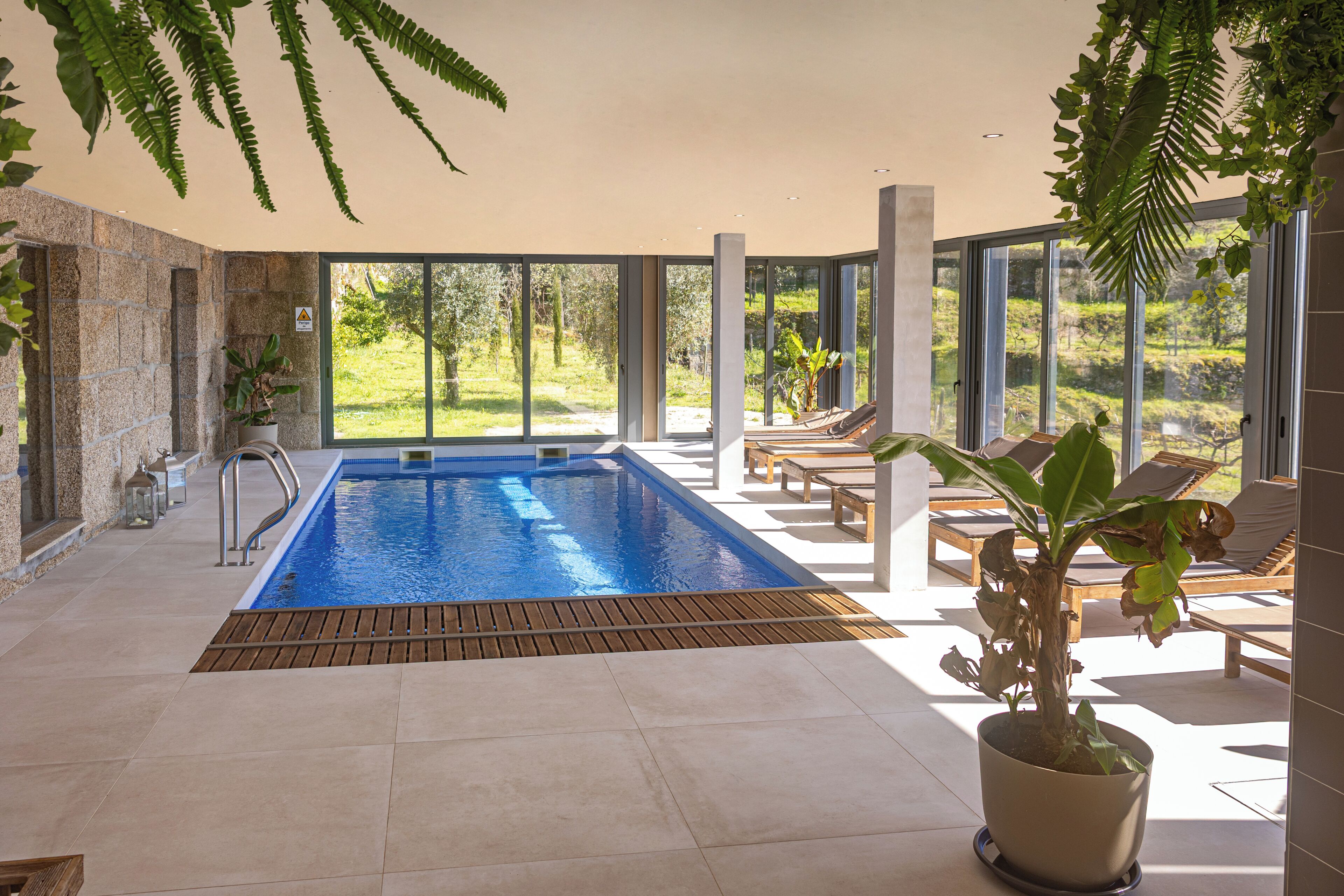 Un área de piscina interior moderna con vistas al exterior, una terraza de madera y sillones de descanso.