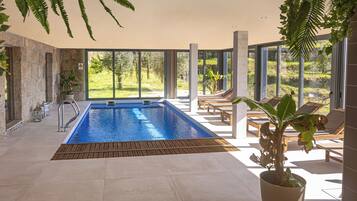 Un área de piscina interior moderna con vistas al exterior, una terraza de madera y sillones de descanso.