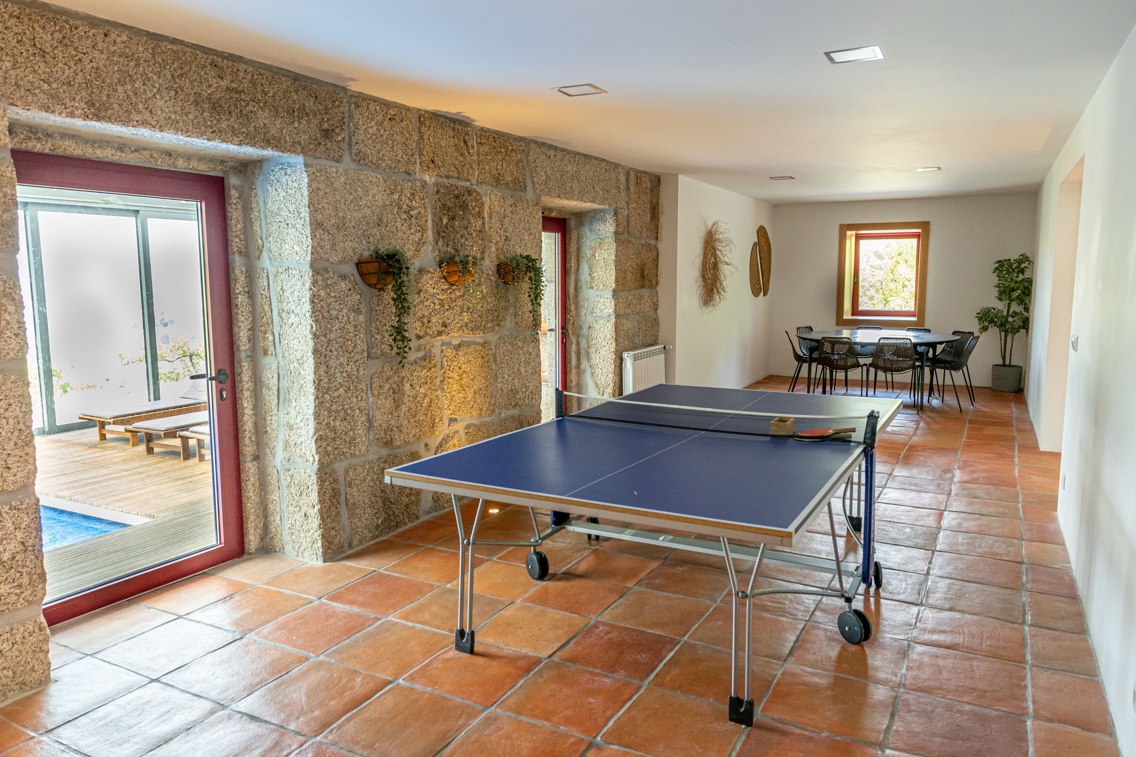 A ping pong table in a room with stone walls, a dining area with chairs, and a view of a pool through a door.